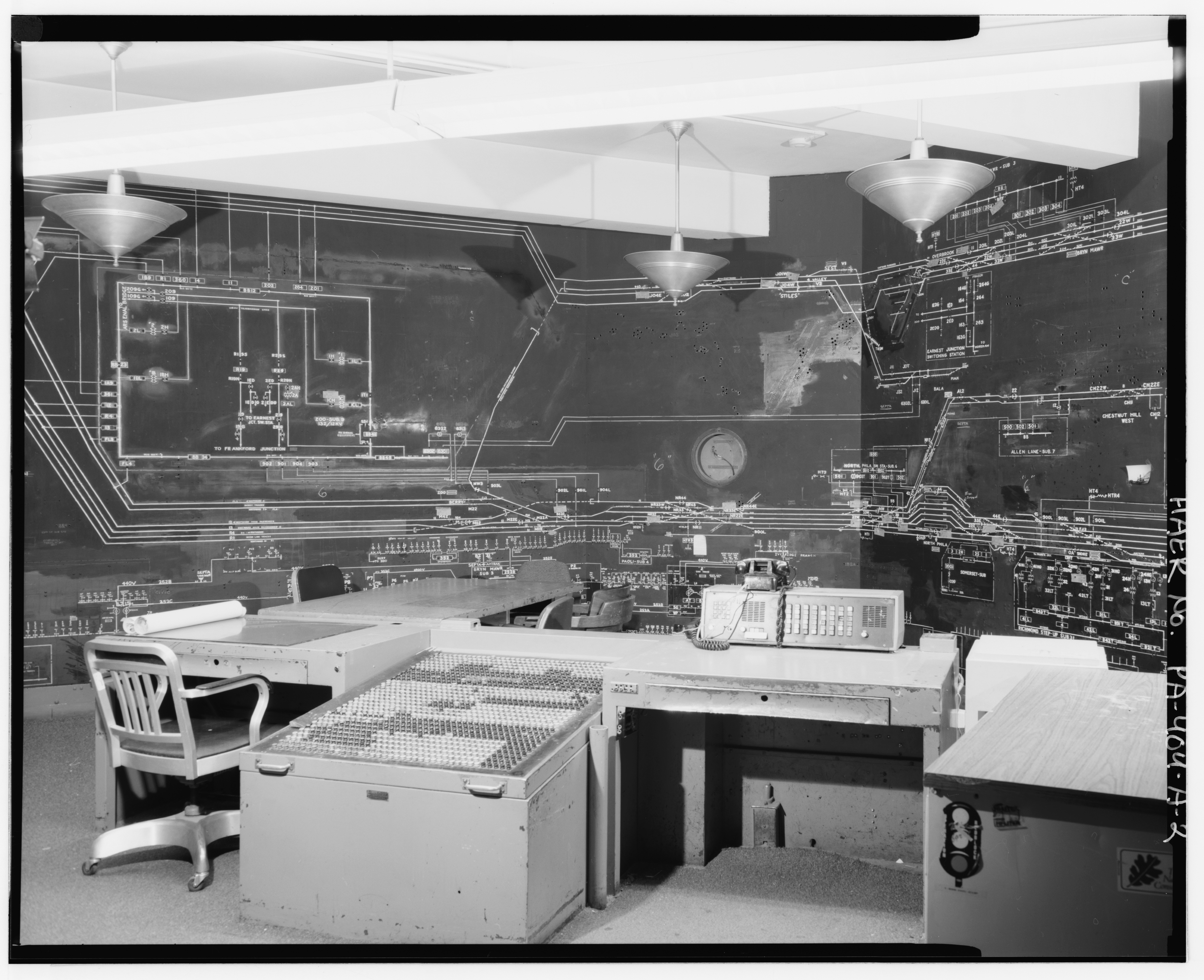 A black-and-white photograph of a control room.
       Pictured in the foreground is a metal desk with a large panel of buttons
       arranged in a grid on the sides.
       In the background is a model board with a track diagram on top and a
       single-line electrical diagram below (mostly hidden by the desk).
       Also on the model board is a circular cutout that once housed a clock.