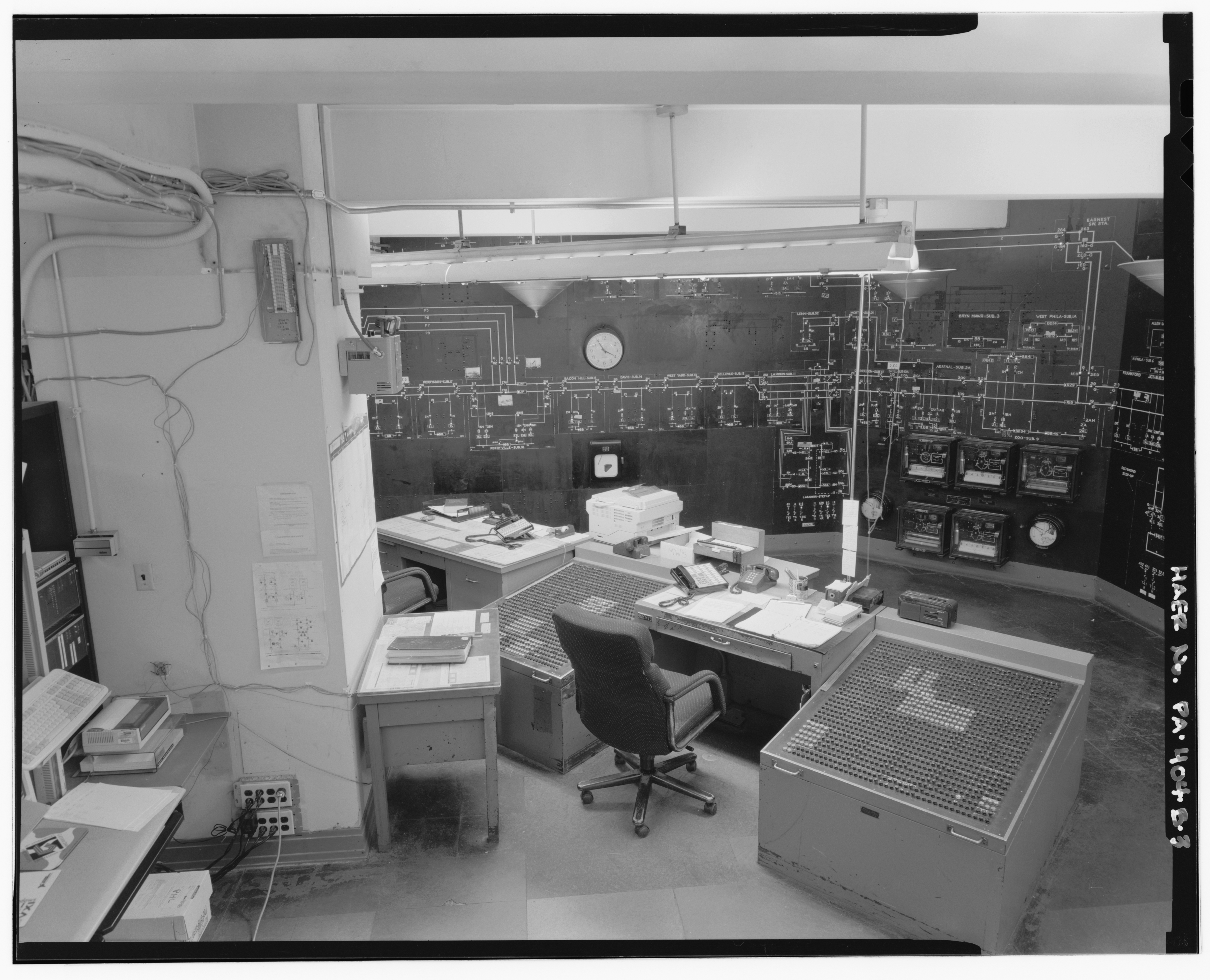 A high-angle black and white photograph of a control room.
       On the left, mostly out of frame, are various computer peripherals,
       a printer, and a rack of telecommunications equipment.
       To the right of that is a large structural column sparsely scattered with
       power plugs and a single 66-type telephone punchdown block mounted on a
       small sheet of plywood.
       To the right of that, in the foreground, is a desk with scattered papers,
       telephones, and a boombox.
       The left and right sides of the desk contain panels of buttons arranged
       in a grid.
       In the background of the desk is a model board with a single-line diagram
       of an electrical network, various pieces of telemetry equipment like
       chart recorders and meters in a cluster, and a clock.